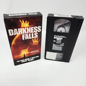 Peut inclure: Une cassette VHS noire et sa boîte pour le film "Darkness Falls". La boîte est principalement rouge et noire, avec le titre du film en grandes lettres blanches et une image enflammée. La cassette est noire avec une étiquette.