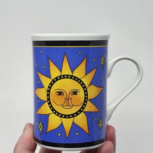 Peut inclure: Un mug en céramique blanche orné d'un fond bleu vif avec un motif central de soleil jaune. Le soleil présente un visage stylisé aux yeux fermés et à l'expression sereine. De petites étoiles jaunes parsèment le fond, le tout encadré par des bandes noires horizontales en haut et en bas.