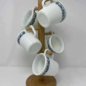 Peut inclure: Un porte-tasses en bois supportant cinq tasses en verre opalin blanc. Chaque tasse présente un motif floral bleu autour du bord. Le support possède une base circulaire et un montant central avec des chevilles en bois pour les tasses.