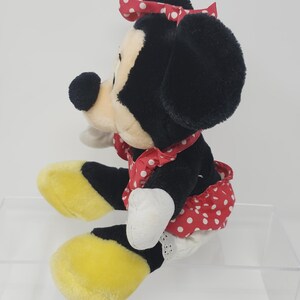 May include: A plush Minnie Mouse toy wearing a red dress with white polka dots and a red bow with white polka dots. The toy has black ears, a black nose, and yellow shoes.