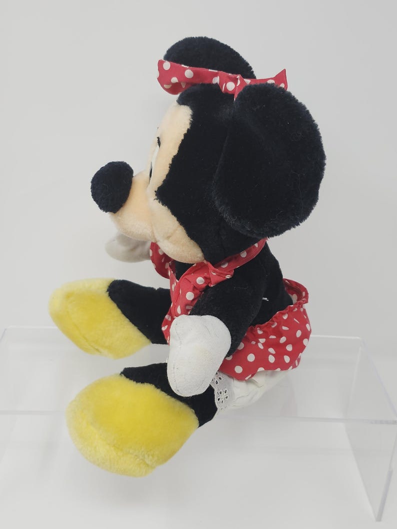 May include: A plush Minnie Mouse toy wearing a red dress with white polka dots and a red bow with white polka dots. The toy has black ears, a black nose, and yellow shoes.
