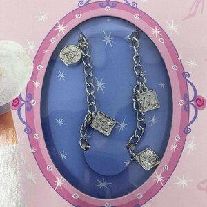 May include: A charm bracelet set in a pink and blue oval-shaped package. The bracelet is silver-toned with multiple charms, including a round charm and square charms. The package has the words "Charm Bracelet" at the top.