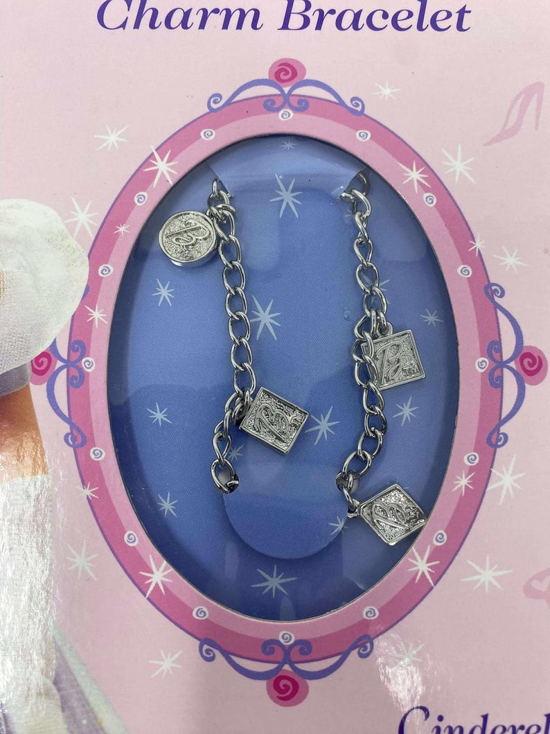 May include: A charm bracelet set in a pink and blue oval-shaped package. The bracelet is silver-toned with multiple charms, including a round charm and square charms. The package has the words "Charm Bracelet" at the top.