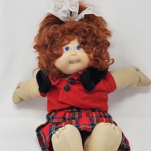 Peut inclure: Une poupée en tissu avec des cheveux roux bouclés attachés par un nœud en dentelle blanche, des yeux bleu vif et une seule dent visible. La poupée porte une veste rouge à revers noirs avec deux boutons noirs et une jupe à carreaux rouge, vert et jaune.