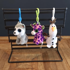 May include: Three plush toy keychains hanging on a black metal rack. The keychain on the left is a grey husky with blue eyes. The keychain in the middle is a purple and pink giraffe with green eyes. The keychain on the right is a white snowman with a black top hat and orange nose.