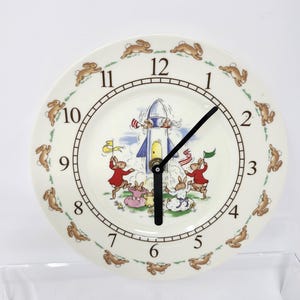 May include: A white ceramic clock with a rocket and bunnies around the edge. The clock face has black hands and brown numbers. The numbers are 1 through 12.
