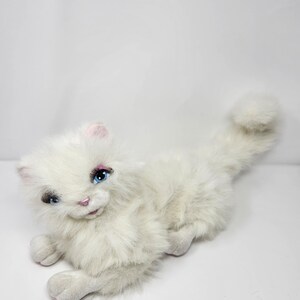 Op de afbeelding: Wit pluche katten speelgoed met blauwe ogen en een roze neus. De kat ligt op zijn buik met zijn staart opgerold.