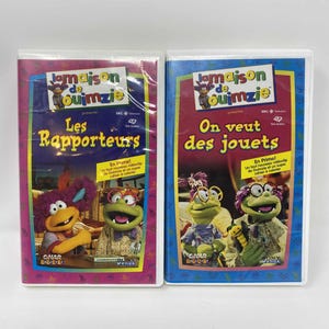 以下が含まれることがあります： 「Jamaison de Ouimzie」というタイトルの2つのVHSテープ。左側のテープは、「Les Rapporteurs」というタイトルと漫画のキャラクターが描かれたピンクと紫色のカバーです。右側のテープは、「On veut des jouets」というタイトルの青いカバーです。