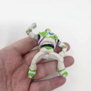 K&ouml;nnte beinhalten: Eine Buzz Lightyear Actionfigur, mit einem wei&szlig;en und gr&uuml;nen Raumanzug mit lila Akzenten. Die Figur ist mit einem erhobenen Arm posiert und zeigt das ikonische Design der Figur aus dem Film Toy Story.
