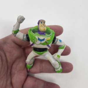 K&ouml;nnte beinhalten: Eine Buzz Lightyear Actionfigur in dynamischer Pose. Das Spielzeug hat einen wei&szlig;en und gr&uuml;nen Raumanzug mit lila Akzenten, einen gr&uuml;nen Helm und einen entschlossenen Gesichtsausdruck. Die Figur wird in einer Hand vor wei&szlig;em Hintergrund gehalten.