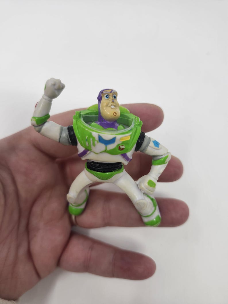 K&ouml;nnte beinhalten: Eine Buzz Lightyear Actionfigur in dynamischer Pose. Das Spielzeug hat einen wei&szlig;en und gr&uuml;nen Raumanzug mit lila Akzenten, einen gr&uuml;nen Helm und einen entschlossenen Gesichtsausdruck. Die Figur wird in einer Hand vor wei&szlig;em Hintergrund gehalten.
