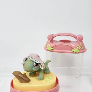 Peut inclure: Figurine de tortue verte et bleue portant un bonnet rose avec de la dentelle blanche, sur une base jaune avec une bûche marron. Un récipient rose avec une poignée et des motifs floraux est à côté de la figurine.