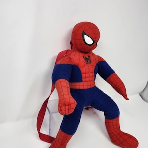 May include: A plush Spider-Man backpack featuring a red and blue fabric body with a black web pattern. The character has large white eyes and a black spider emblem on the chest. Red fabric straps are attached to the back for carrying purposes.