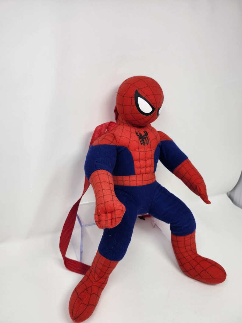 May include: A plush Spider-Man backpack featuring a red and blue fabric body with a black web pattern. The character has large white eyes and a black spider emblem on the chest. Red fabric straps are attached to the back for carrying purposes.