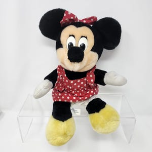 May include: A plush black and white Minnie Mouse toy wearing a red dress with white polka dots and a red bow. The toy is sitting on a clear acrylic shelf.