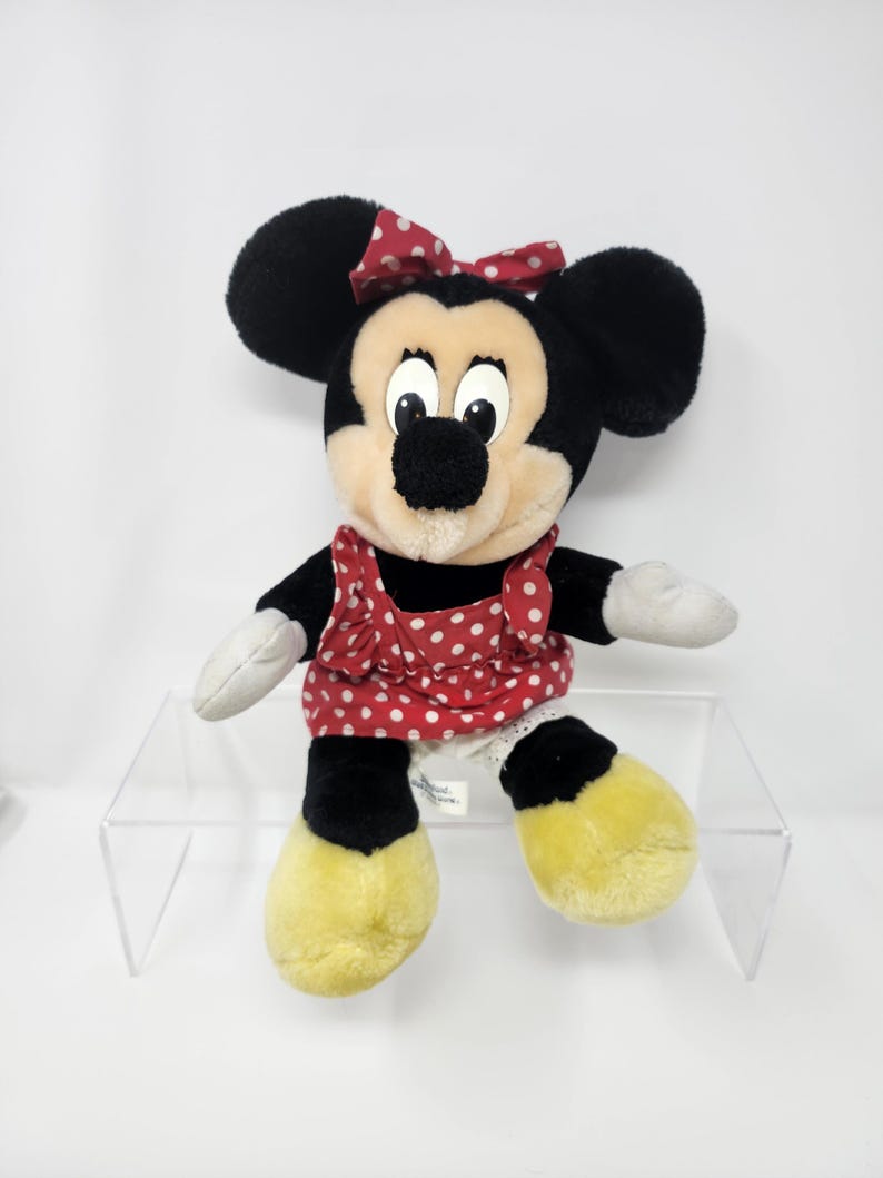 May include: A plush black and white Minnie Mouse toy wearing a red dress with white polka dots and a red bow. The toy is sitting on a clear acrylic shelf.