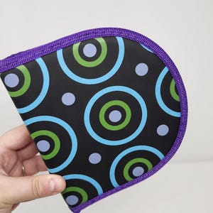 May include: A semi-circular case featuring a black background with a repeating pattern of concentric circles in light blue, lime green, and lavender. The edges of the case are finished with a textured purple fabric trim along the curved border.