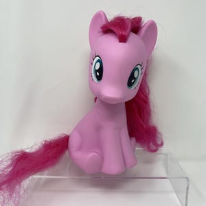 May include: A pink toy pony with a magenta mane and tail. The pony has large blue eyes with white highlights. The toy is sitting on a clear plastic display stand. The pony's body is a solid pink color.