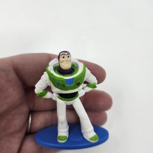 K&ouml;nnte beinhalten: Eine kleine Buzz Lightyear-Figur, die einen wei&szlig;en und gr&uuml;nen Raumanzug mit lila Akzenten tr&auml;gt. Das Spielzeug steht auf einer blauen Basis, die Arme in die H&uuml;ften gestemmt. Ein Sammlerst&uuml;ck aus der Toy Story-Filmreihe.