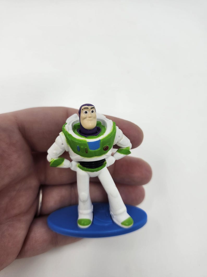 K&ouml;nnte beinhalten: Eine kleine Buzz Lightyear-Figur, die einen wei&szlig;en und gr&uuml;nen Raumanzug mit lila Akzenten tr&auml;gt. Das Spielzeug steht auf einer blauen Basis, die Arme in die H&uuml;ften gestemmt. Ein Sammlerst&uuml;ck aus der Toy Story-Filmreihe.