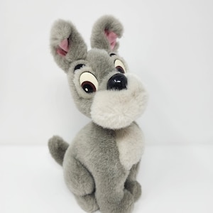 Può includere: Un peluche grigio a forma di cane, con grandi occhi espressivi e orecchie flosce. Il cane ha un muso e un petto grigio chiaro, un naso nero e una piccola coda ricurva. Il giocattolo è seduto, con la testa leggermente inclinata.