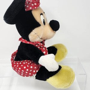 May include: A plush toy of Minnie Mouse wearing a red and white polka dot dress and a red headband with white polka dots. The toy is sitting with its legs crossed and has a yellow and white plush face.