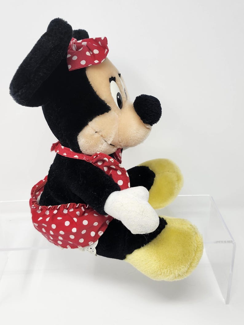 May include: A plush toy of Minnie Mouse wearing a red and white polka dot dress and a red headband with white polka dots. The toy is sitting with its legs crossed and has a yellow and white plush face.
