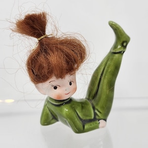 Puede incluir: Figura de cerámica de un niño con un traje verde y un moño rojo. La figura está tumbada boca arriba con una pierna levantada. La cara tiene rasgos pintados, incluyendo mejillas rosadas y una pequeña sonrisa. Es una pieza de colección vintage.