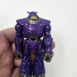 K&ouml;nnte beinhalten: Eine lila Actionfigur eines roboterartigen Charakters mit einem bedrohlichen Gesichtsausdruck. Die Figur hat ein detailliertes Design mit metallischem Aussehen, einem lila K&ouml;rper, grauen Akzenten und einem gelben Mund. Das Spielzeug steht aufrecht.