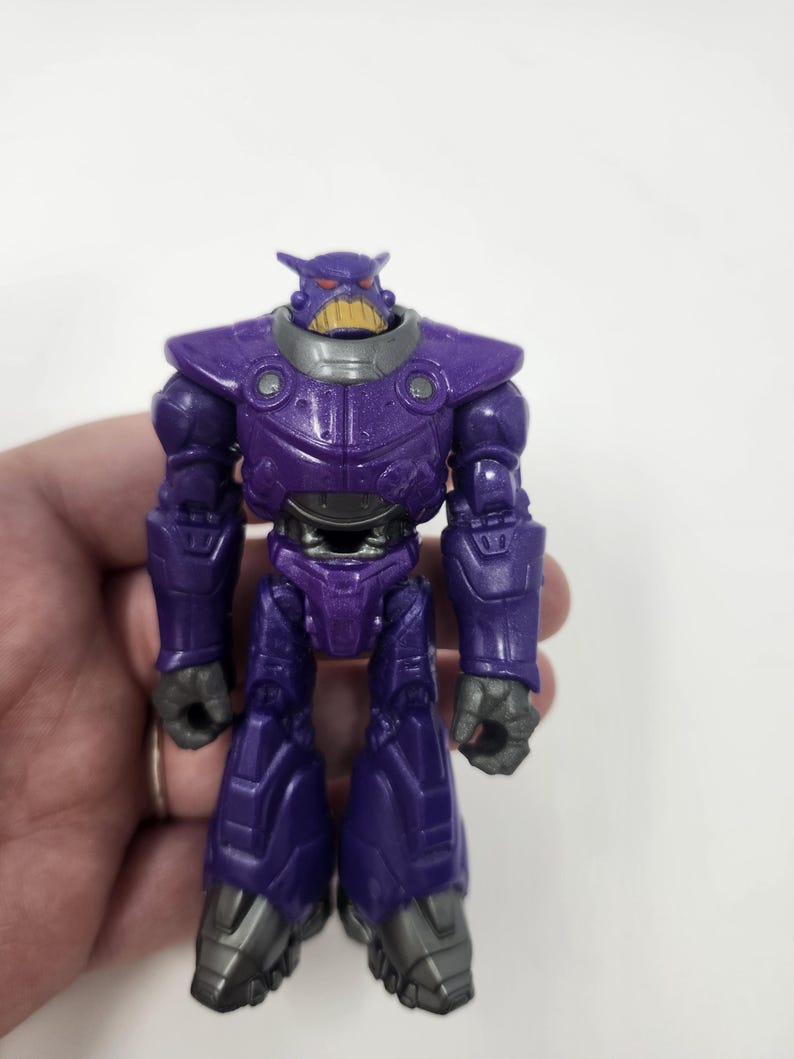K&ouml;nnte beinhalten: Eine lila Actionfigur eines roboterartigen Charakters mit einem bedrohlichen Gesichtsausdruck. Die Figur hat ein detailliertes Design mit metallischem Aussehen, einem lila K&ouml;rper, grauen Akzenten und einem gelben Mund. Das Spielzeug steht aufrecht.
