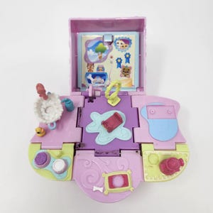 Puede incluir: Un juego de juguetes vintage rosa y morado abierto, con varios compartimentos y accesorios. El juego incluye una pequeña imagen, una cama, un hueso de perro y otros objetos en miniatura. El juguete está diseñado para el juego imaginativo.