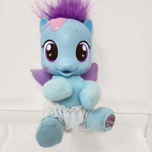 Könnte beinhalten: Hellblaues My Little Pony Plüschtier mit lila Haaren und Flügeln. Das Spielzeug hat große Augen mit lila Akzenten, eine rosa Markierung auf dem Kopf und trägt eine weiße Windel. Das My Little Pony-Logo befindet sich auf dem Fuß.