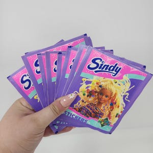 Könnte beinhalten: Eine Hand hält einen Fächer aus lila Sindy-Aufkleberpackungen. Jede Packung hat eine rosa Oberseite mit dem Sindy-Logo und einem Bild einer blonden Puppe. Das Wort "STICKERS" ist unten auf jeder Packung aufgedruckt.