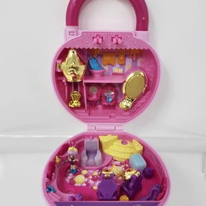 Può includere: Un playset giocattolo rosa a forma di cuore, aperto per rivelare un interno in miniatura. Il set presenta una maniglia, una vanità color oro e piccole statuette. L'interno è decorato in tonalità rosa e viola.