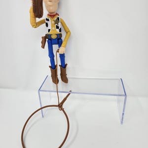 K&ouml;nnte beinhalten: Woody-Actionfigur, bekleidet mit einem gelben Hemd, blauen Hosen und braunen Stiefeln, h&auml;lt einen braunen Hut. Die Figur hat ein Sheriff-Abzeichen und einen braunen Lasso. Das Spielzeug steht auf einem transparenten Acryl-Displayst&auml;nder.