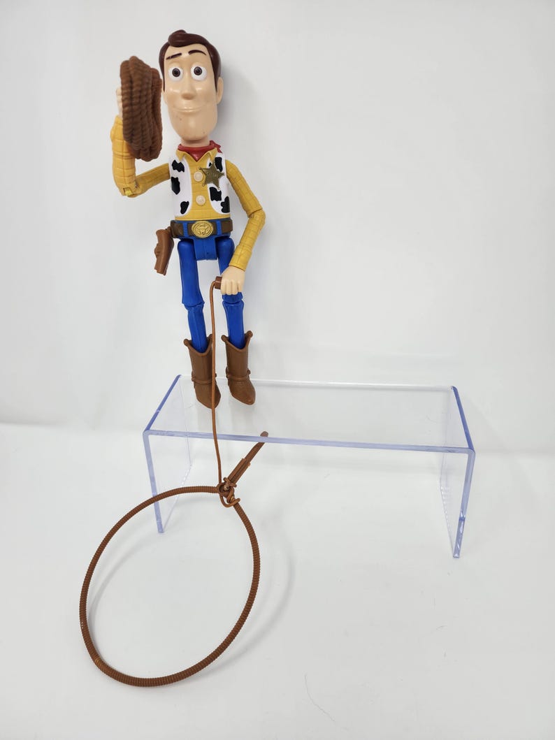K&ouml;nnte beinhalten: Woody-Actionfigur, bekleidet mit einem gelben Hemd, blauen Hosen und braunen Stiefeln, h&auml;lt einen braunen Hut. Die Figur hat ein Sheriff-Abzeichen und einen braunen Lasso. Das Spielzeug steht auf einem transparenten Acryl-Displayst&auml;nder.