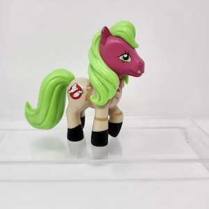 May include: A toy pony figurine with a pink head, green mane and tail, and a tan body. The pony has black hooves and a Ghostbusters logo on its side. The figurine is on a clear, reflective surface.