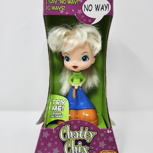 Peut inclure: Une poupée Chatty Chix aux cheveux blonds, portant un haut à col roulé vert, un pantalon bleu et des chaussures orange, emballée dans une boîte violette avec le texte I SAY NO WAY! 10 WAYS!, NO WAY!, TRY ME! Bobble my head! et Chatty Chix.