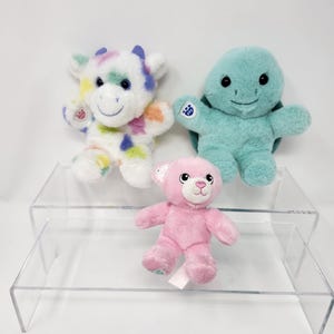 Peut inclure: Trois peluches sur une étagère en acrylique transparente. Les peluches sont un ours rose, une tortue verte et une vache blanche avec des taches multicolores.