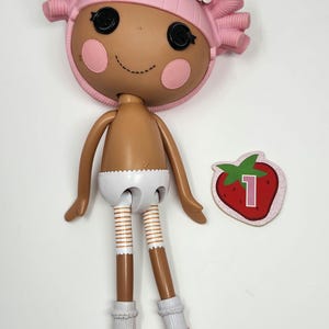 Puede incluir: Una muñeca Lalaloopsy con cabeza rosa y pelo rizado, ojos negros de botón y mejillas rosadas. La muñeca lleva un pañal blanco, zapatos naranjas y calcetines blancos. Una tarjeta con el número uno en forma de fresa está al lado de la muñeca.