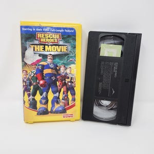 Peut inclure: Une cassette VHS jaune et une cassette VHS noire. L'étui présente le titre "Rescue Heroes: The Movie" avec des personnages de dessins animés. Le texte "Starring in their FIRST Full-Length Feature!" est également visible.