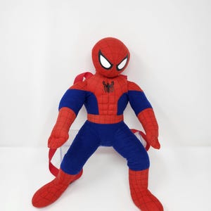 May include: A plush Spider-Man backpack featuring a red and blue fabric design with a black spider emblem on the chest. The character has a round head with white eye patches and a web pattern on the red sections, including red adjustable straps.