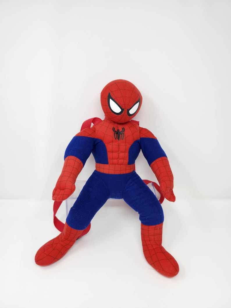 May include: A plush Spider-Man backpack featuring a red and blue fabric design with a black spider emblem on the chest. The character has a round head with white eye patches and a web pattern on the red sections, including red adjustable straps.