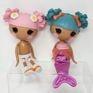Puede incluir: Dos muñecas Lalaloopsy. Una muñeca tiene el pelo rosa con detalles de flores blancas, y la otra tiene el pelo verde azulado con lazos naranjas. Una muñeca lleva una cola de sirena, y la otra calcetines blancos y zapatos naranjas.