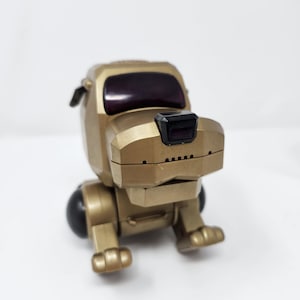 May include: A gold-coloured robotic dog toy with a blocky, geometric design. It features a dark, rectangular visor for eyes, a small black nose, and rounded black joints at the shoulders. The figure is depicted in a seated position with a smooth finish.