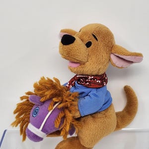 以下が含まれることがあります： 青いシャツと赤いバンダナを着た、茶色のカンガルーのようなキャラクターのぬいぐるみ。キャラクターは茶色のたてがみを持つ紫色の馬に乗っています。おもちゃは白い背景にあります。