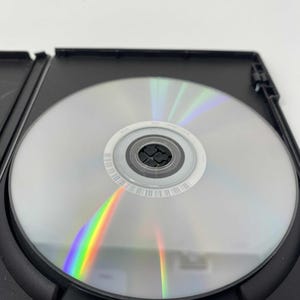 May include: A silver DVD disc inside a black plastic case. The disc reflects a rainbow of colors. The case is open, revealing the disc's surface and the surrounding black plastic.