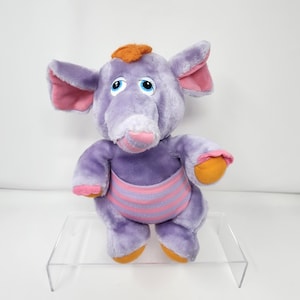 Op de afbeelding: Een pluche paarse olifant met grote oren, blauwe ogen en een roze en oranje gestreepte buik. De olifant heeft oranje accenten op zijn voeten, handen en een pluk haar. Het speelgoed staat op een heldere acryl standaard.