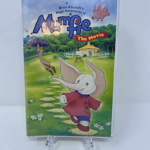 Puede incluir: Una cinta VHS de "Mumfie The Movie". La portada muestra un elefante de dibujos animados con un abrigo rojo, un paisaje verde, un cerdo volador y el título en azul y amarillo.