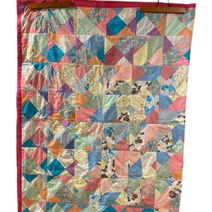 May include: A rectangular patchwork quilt featuring a grid of squares, each composed of smaller triangles in various pastel colours including pink, blue, yellow, and green. Some squares contain floral or small toy patterns, all bordered by a solid pink trim.
